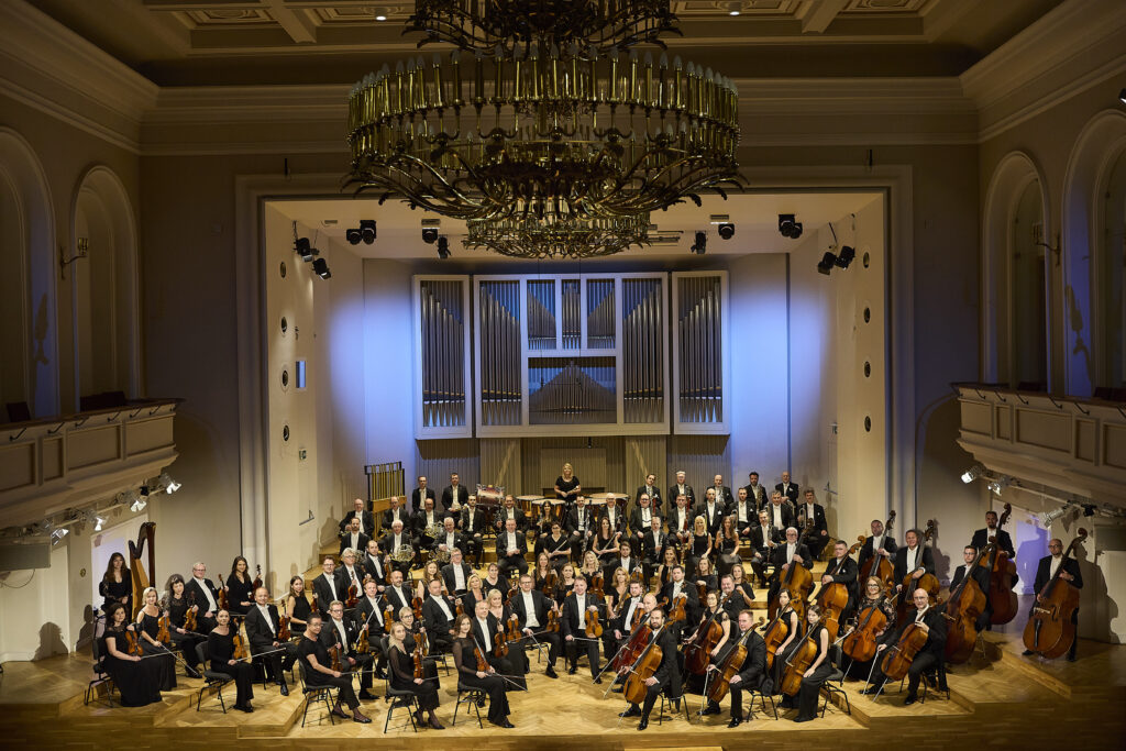 Orkiestra Symfoniczna Filharmonii Śląskiej siedzi na estradzie w składzie około 60 osób, muzycy są ubrani w czarne stroje i trzymają w rękach instrumenty