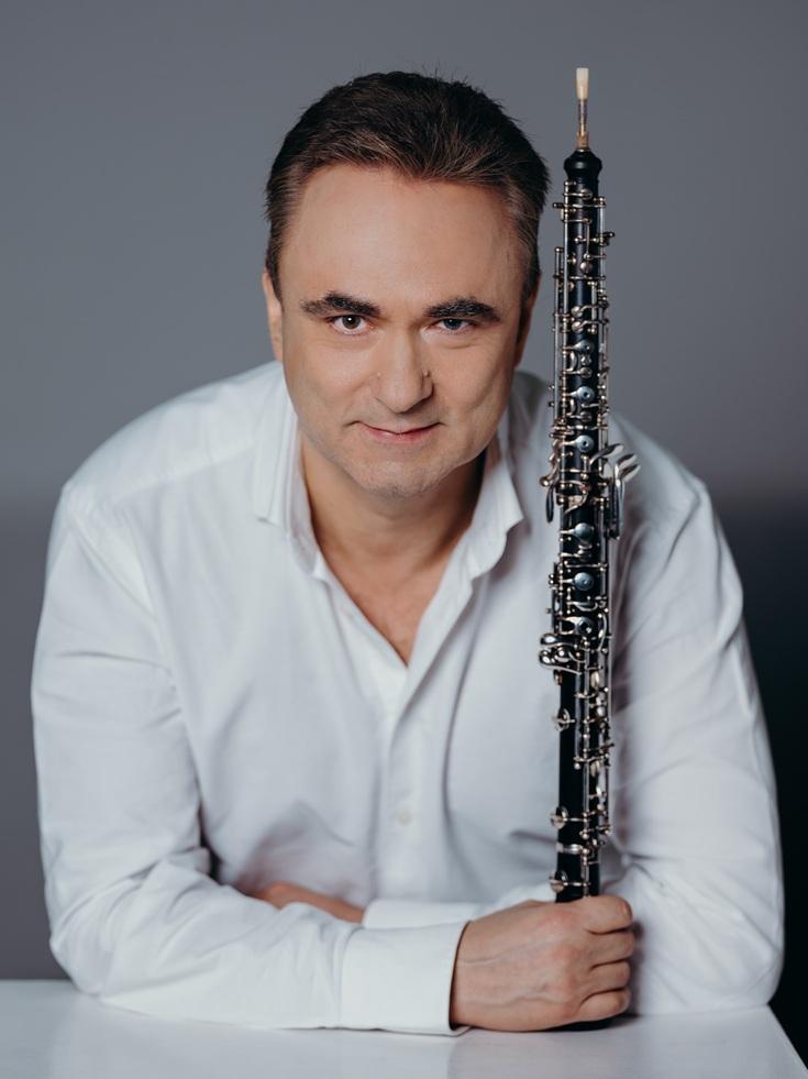 Oboista pozuje ze swoim czarnym, długim i drewnianym instrumentem, ubrany jest w białą koszulę