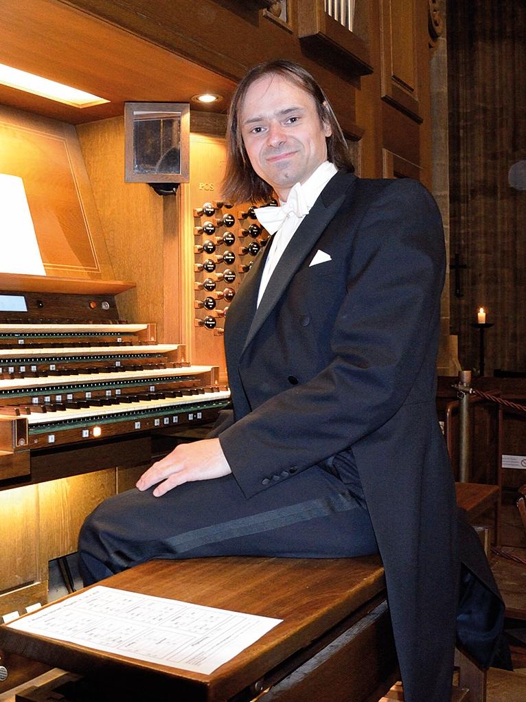 Organista Tomasz Orlow siedzi przed klawiaturą organów, jest ubrany w czarny smoking oraz białą koszulę i białą muszkę